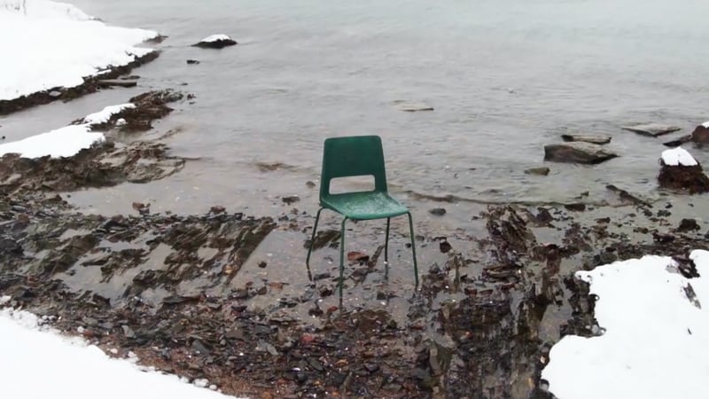 En grønn stol i plastikk står i vannkanten med snødekt landskap